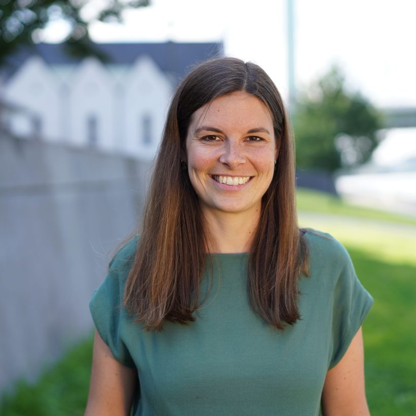 Anna Reichert, Referentin des Community Resilience Lab, lächelt in grüner Bluse vor einer Grünanlage am Flussufer.