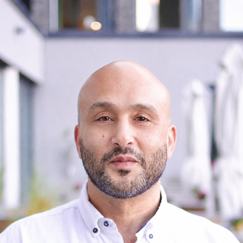 Hicham Boutouil, Referent des Community Resilience Lab, lächelt mit kurzem Bart in weißem Hemd vor unscharfem Hintergrund.
