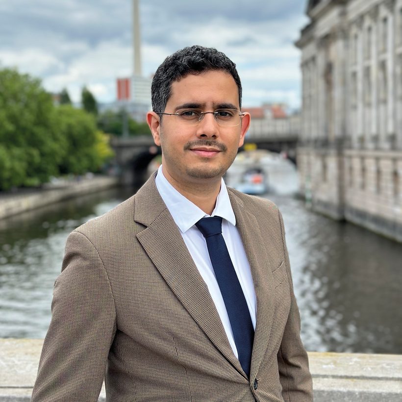Mimoun Berrissoun, Referent des Community Resilience Lab, lächelt in beigem Sakko mit weißem Hemd und dunkler Krawatte vor einem Fluss mit historischem Gebäude.
