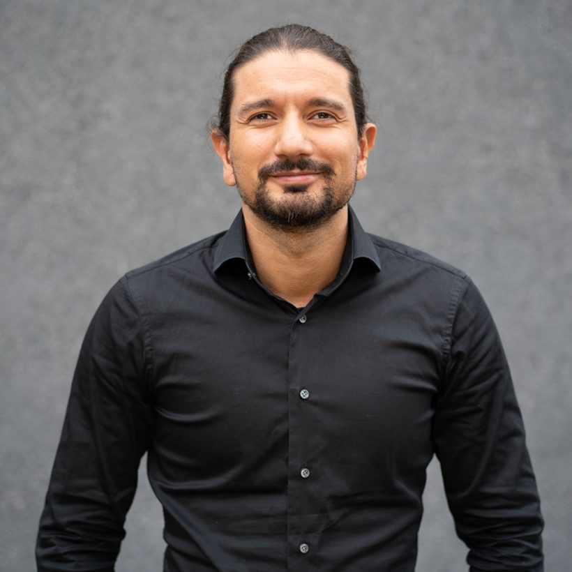 Numan Özer, Referent des Community Resilience Lab, mit Vollbart und zusammengebundenem Haar in schwarzem Hemd vor einer grauen Wand.